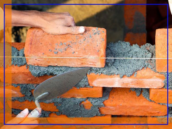 A close-up of hands laying bricks with mortar on a construction site, showing bricklaying in progress.