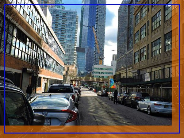 A street in Queens lined with parked cars and a mix of residential and commercial buildings, with a tall building under construction in the distance.